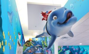 A friendly dolphin and crab race through a dental office hallway with faux coral archways and themed murals