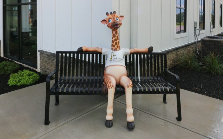 Young giraffe mascot sitting on an exterior bench outside a dental office