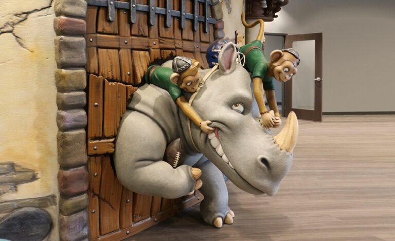 side view of sculpted football playing rhino charging through castle door with monkeys on his back
