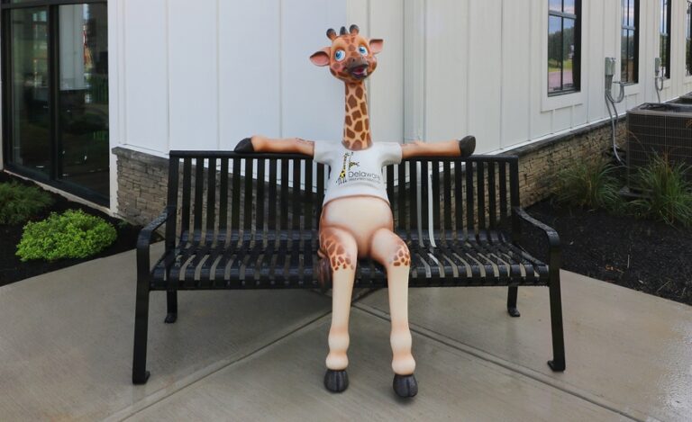 A cool teenage giraffe mascot sitting on an exterior bench, waiting to take a a photo.