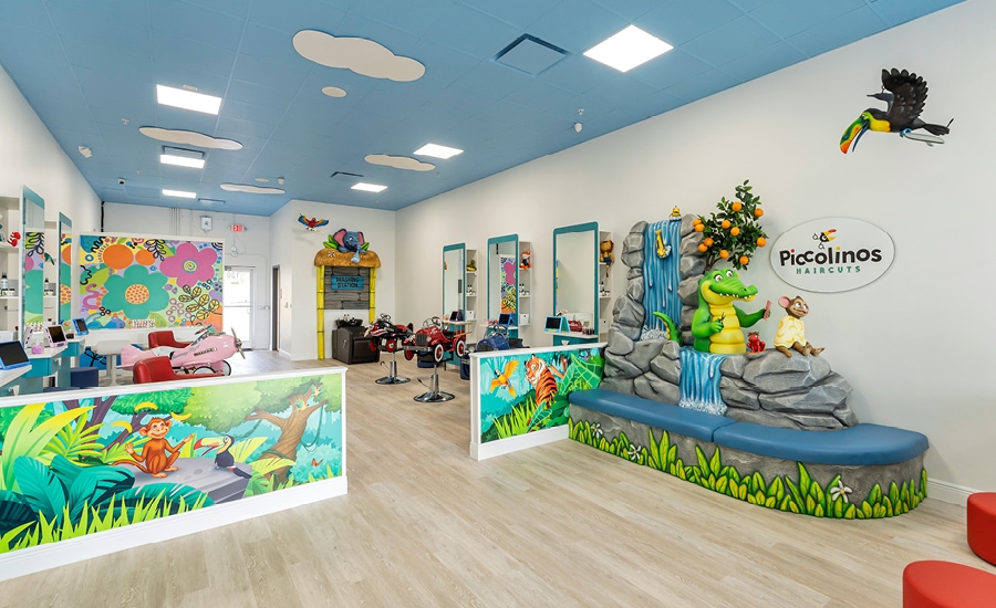 Full view of jungle-themed kid-friendly hair salon showing partition walls with 2D murals, a washing station with 3D sculpted sign, and large waiting area bench wrapped around a 3D sculpted waterfall and characters